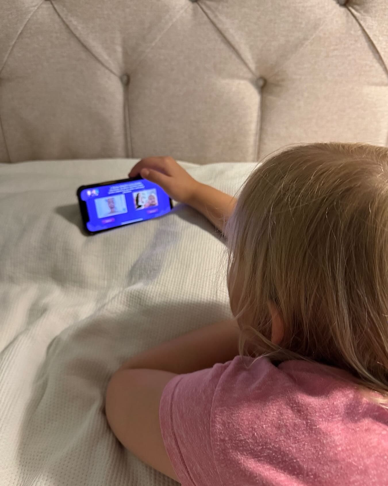Lapsi sängyllä pelaa ÄrräTreeni-sovellusta puhelimella, pinkki paita. Kuva otettu takaviistosta, anonyymi mutta lämmin.
